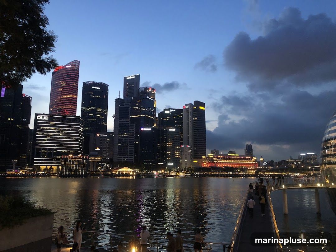 A wide shot of a city skyline at dusk, with tall buildings illuminated by lights. A body of water reflects the city lights, and a walkway with people can be seen in the foreground. The sky is a deep blue with some clouds.