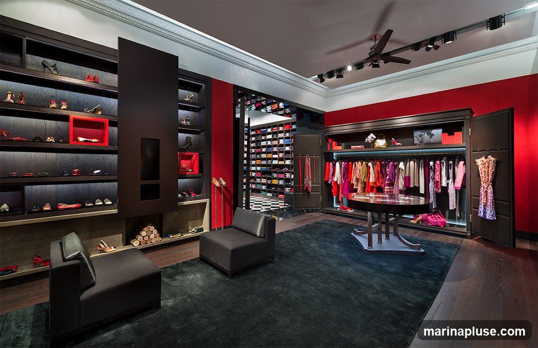 A high-end boutique interior featuring a display of women's shoes on shelves, a rack of colorful clothing, and a central display table. The store has dark wood shelving, red accent walls, and plush carpeting.
