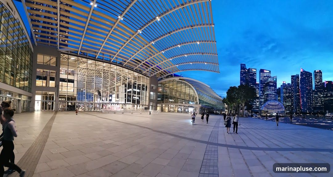 A wide-angle view of a modern architectural complex with a distinctive curved roof structure, illuminated at dusk. The building features extensive glass facades, revealing interior spaces. In the background, a vibrant city skyline with numerous skyscrapers is visible against a deep blue sky. Several people are walking in the plaza area in front of the building.