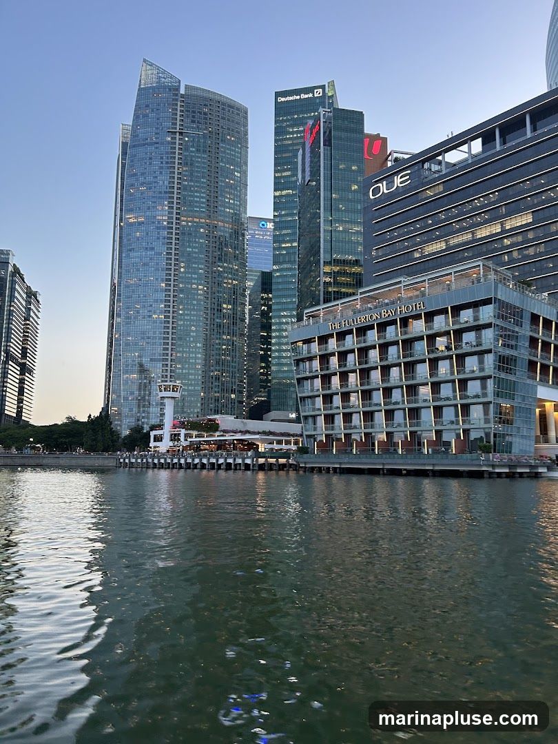 A wide shot of a city skyline at dusk, with a body of water in the foreground. Several modern skyscrapers dominate the background, including buildings with signage for Deutsche Bank and OUE. In the mid-ground, The Fullerton Bay Hotel is prominently visible, a multi-story building with a distinctive facade and balconies. The water reflects the lights of the city and the buildings.