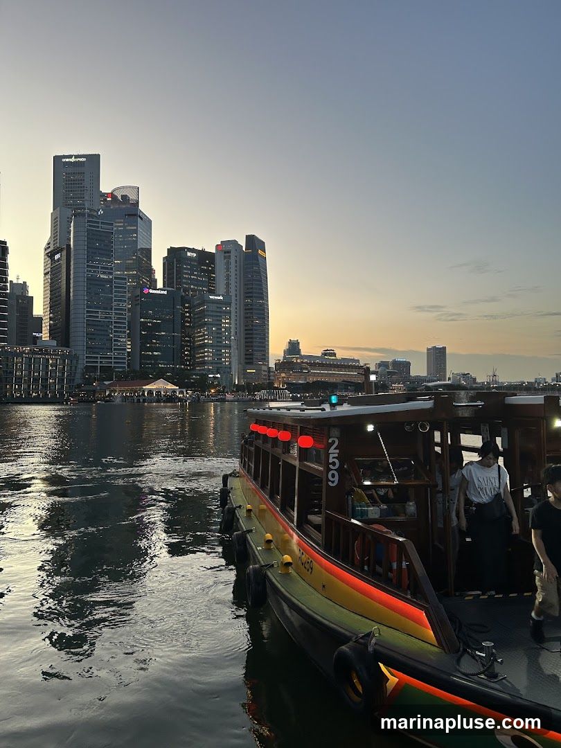 A boat with red lanterns and yellow and green accents is docked on the water. People are visible on the deck of the boat. In the background, a city skyline with numerous skyscrapers is illuminated against a dusky sky. The water reflects the city lights and the boat.