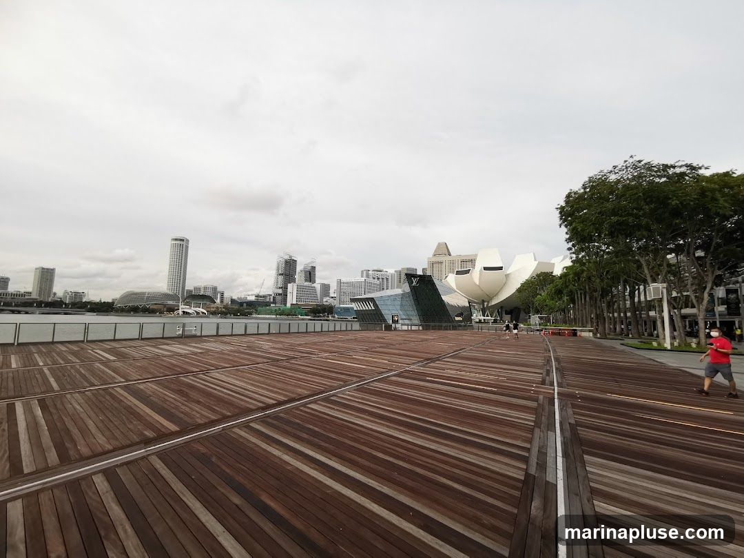 A wide wooden promenade with a view of the Marina Bay Sands complex and the Singapore skyline under a cloudy sky. Several people are visible walking on the promenade.