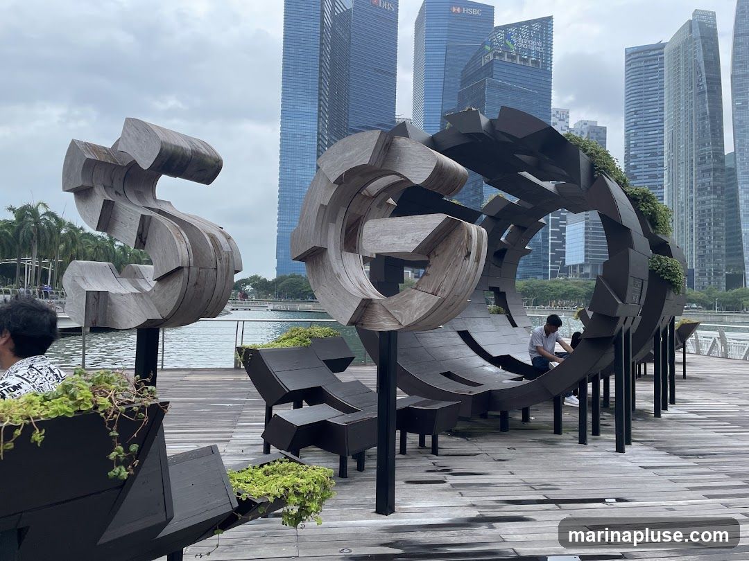 A waterfront area featuring large wooden sculptures and modern seating. In the foreground, a person is seated within a large, circular, segmented sculpture adorned with greenery. To the left, another abstract wooden sculpture resembling a stylized letter 'S' stands prominently. The area is paved with wooden planks, and in the background, a cityscape with tall skyscrapers is visible under an overcast sky.