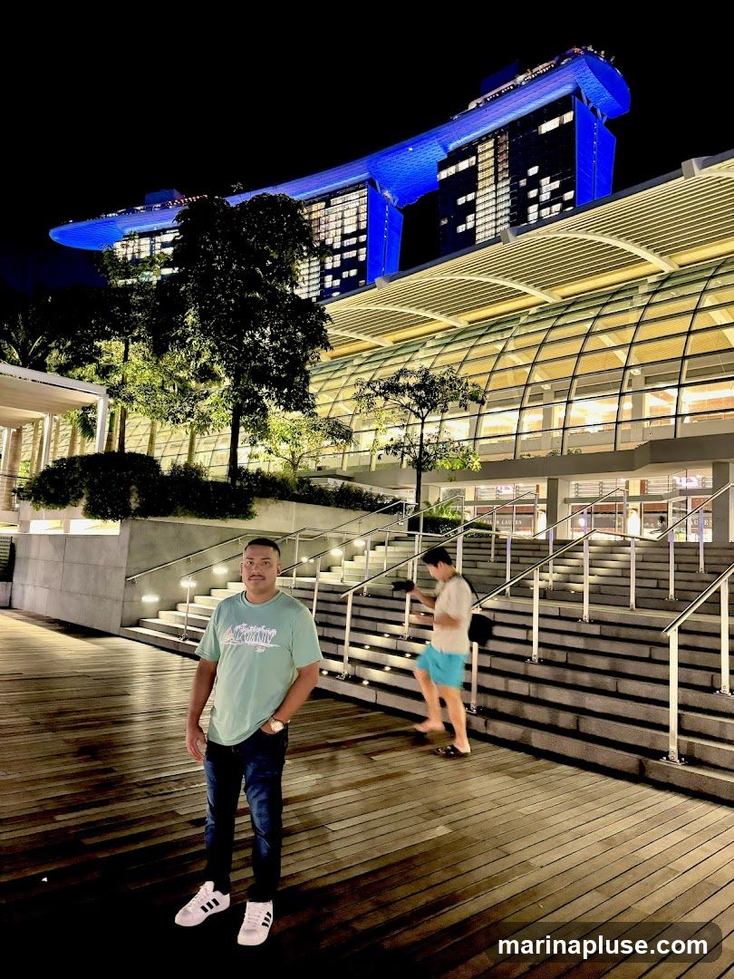 A man stands on a wooden walkway at night, with a large building illuminated in blue in the background. Another person is walking up a set of stairs to the right. The architecture suggests a modern urban setting.