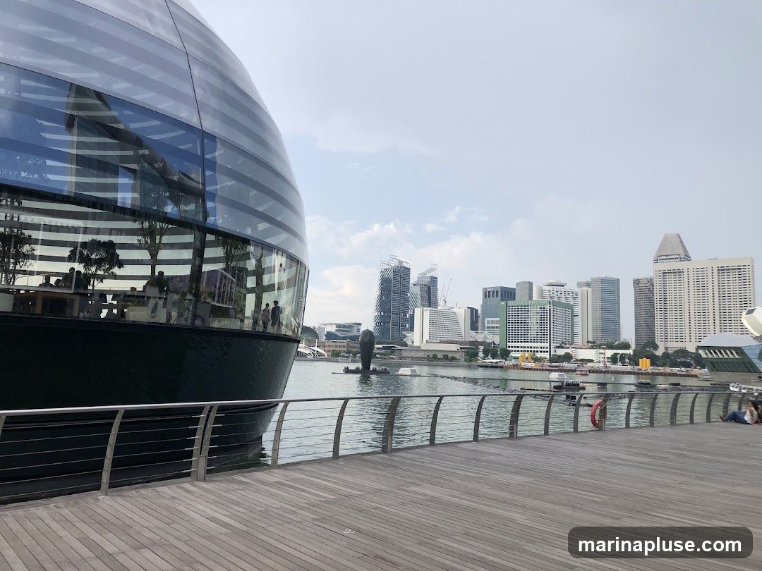 A large, spherical glass building with horizontal banding is reflected in the water. Inside, people can be seen sitting at tables and walking around. In the background, a cityscape with numerous tall buildings rises against a cloudy sky. A wooden boardwalk with a metal railing runs along the foreground, with a few people seated on it.