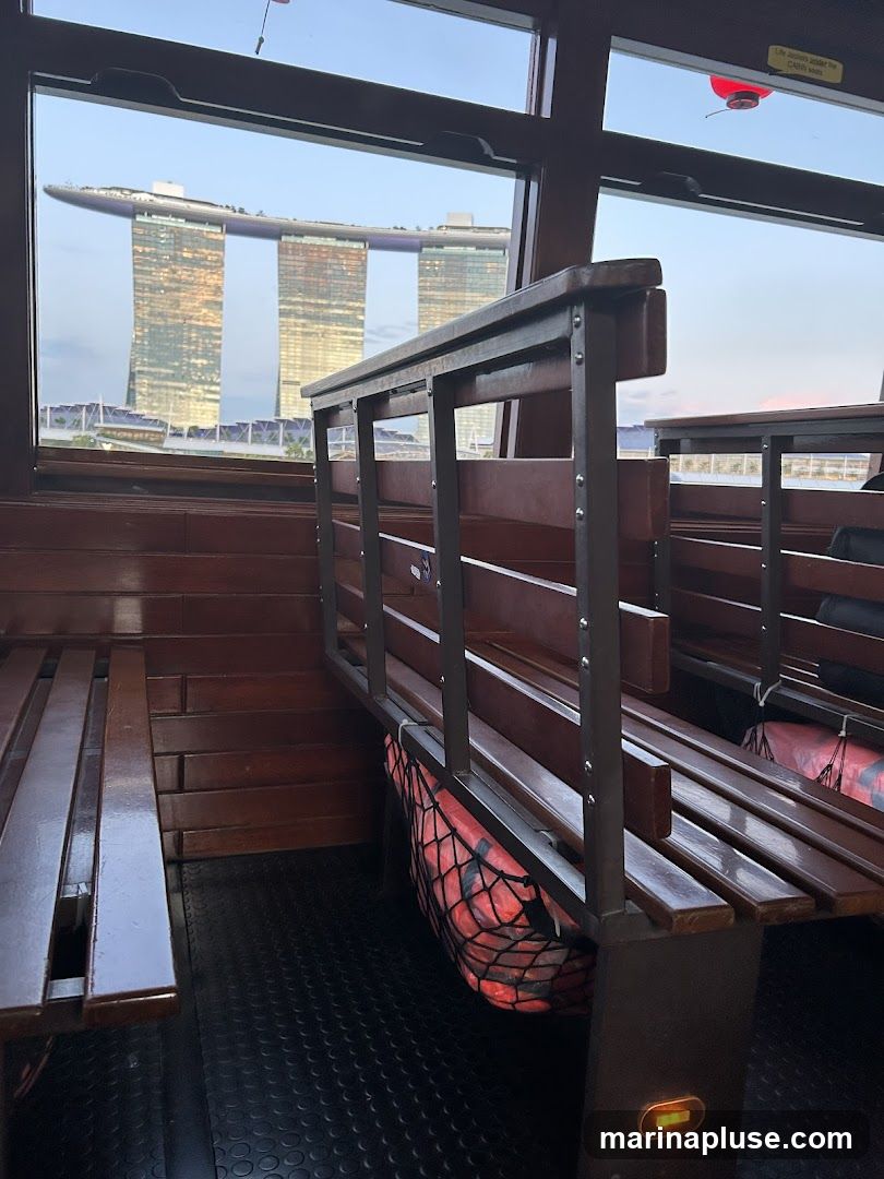 The image shows a view of the Marina Bay Sands hotel in Singapore from inside a boat. The boat has wooden benches and railings. A life vest is visible under one of the benches. The sky is clear and the sun is setting, casting a warm glow on the hotel.