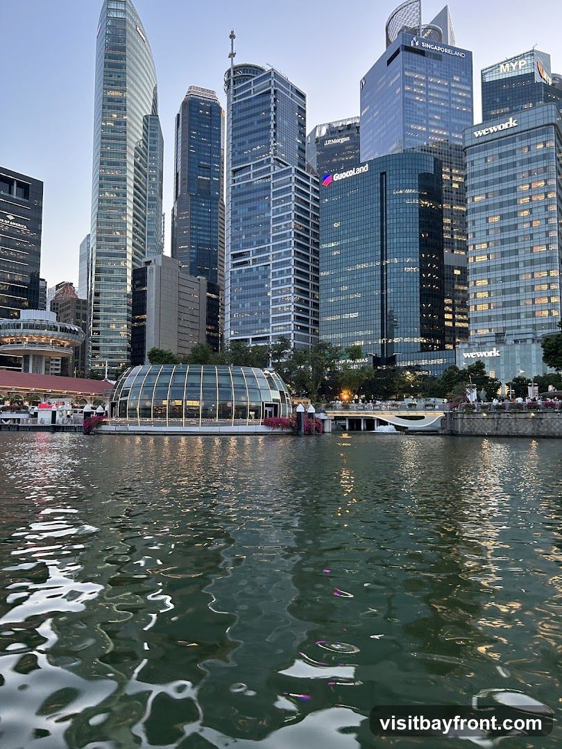 A view of a modern cityscape with numerous skyscrapers and a distinctive glass-domed building situated by a body of water. The water in the foreground reflects the lights and structures of the city. The sky is clear and blue, suggesting it is either early morning or late afternoon. Signs for various corporations like J.P. Morgan, GuocoLand, and WeWork are visible on the buildings.