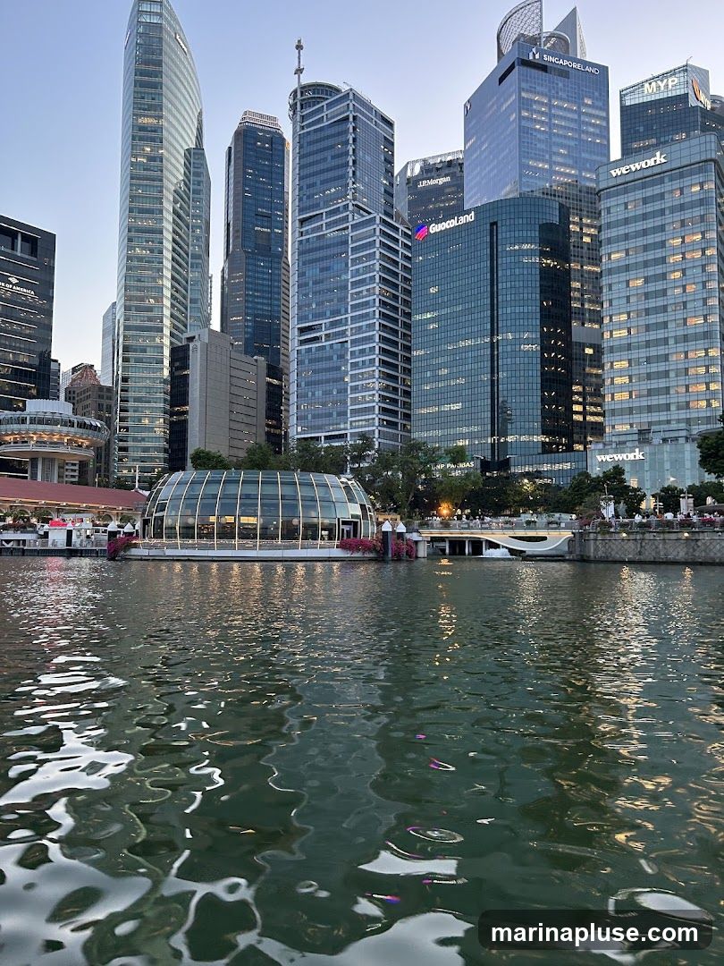 A view of a modern cityscape with numerous skyscrapers and a distinctive glass-domed building situated by a body of water. The water in the foreground reflects the lights and structures of the city. The sky is clear and blue, suggesting it is either early morning or late afternoon. Signs for various corporations like J.P. Morgan, GuocoLand, and WeWork are visible on the buildings.