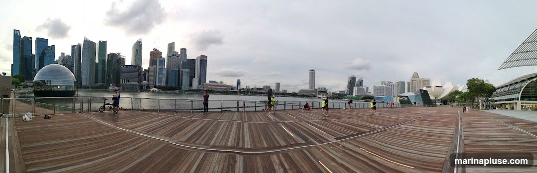 A panoramic view of the Singapore skyline from a wooden boardwalk. The iconic Marina Bay Sands complex, including the lotus-shaped ArtScience Museum and the Apple Store, is visible across the water. Several skyscrapers form the city's financial district. People are seen walking and exercising on the boardwalk. The sky is overcast.