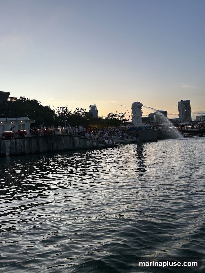 A wide shot of the Merlion fountain in Singapore, with its iconic statue spouting water. The background shows a city skyline with tall buildings and trees, silhouetted against a dusky sky. A body of water, likely a river or bay, fills the foreground, reflecting the sky and structures. A crowd of people is gathered on the waterfront promenade, observing the fountain and the cityscape. The overall scene captures a popular tourist spot during the evening hours.