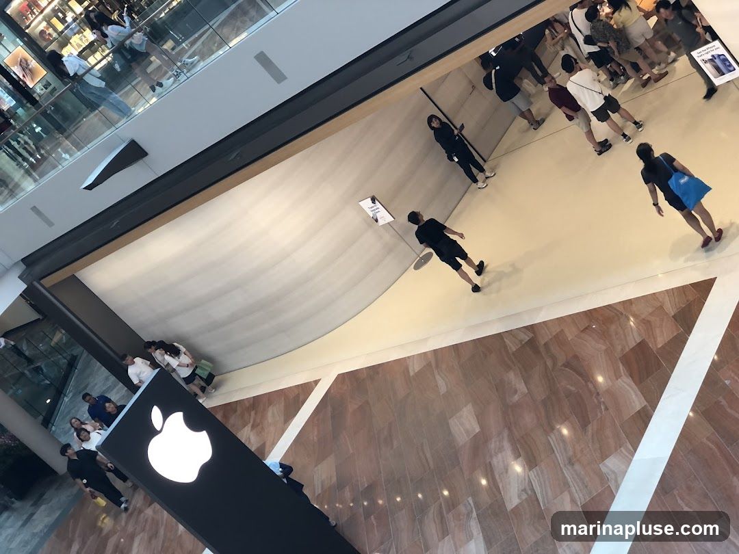 An overhead view of the interior of an Apple Store in a mall. People are browsing and interacting with displays. A large illuminated Apple logo is prominently displayed.