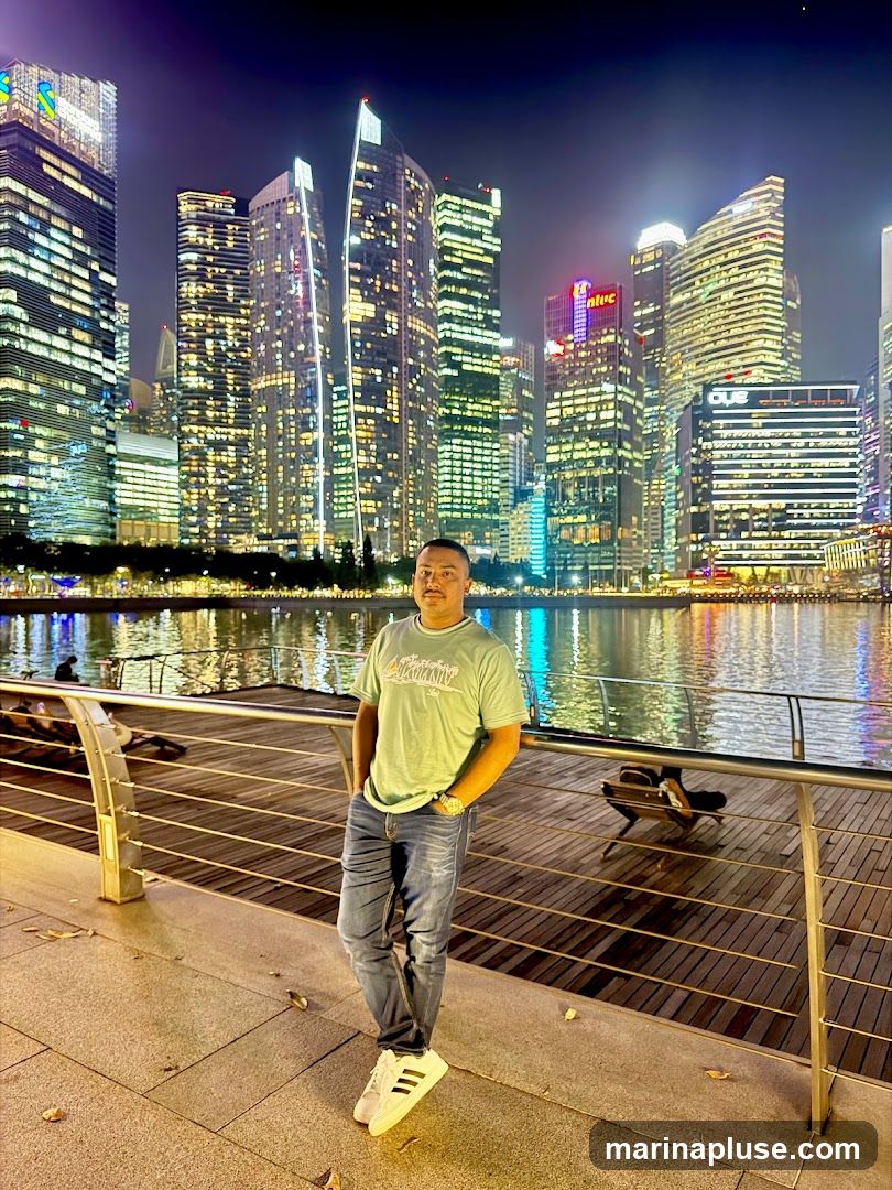 A man stands on a wooden boardwalk, posing for a photo with a backdrop of a brightly lit city skyline at night. The skyscrapers are illuminated with countless windows, reflecting in the water in front of them. The man is wearing a light green t-shirt, blue jeans, and white sneakers. The boardwalk has a metal railing and wooden planks.