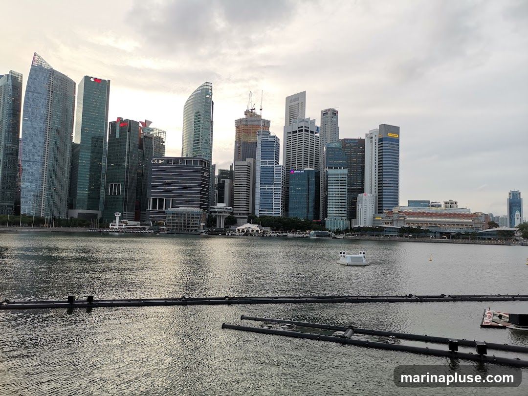 A panoramic view of the Singapore skyline with modern skyscrapers and a body of water in the foreground. The sky is overcast.