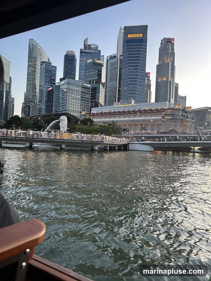 A view of the Singapore skyline featuring numerous skyscrapers, including the Maybank building and the Fullerton Hotel. The iconic Merlion statue is visible in the foreground, with people gathered along the waterfront. The image is taken from a boat on the water, with the edge of the boat visible in the lower left corner. The sky is clear and the sun is setting, casting a warm glow on the buildings.