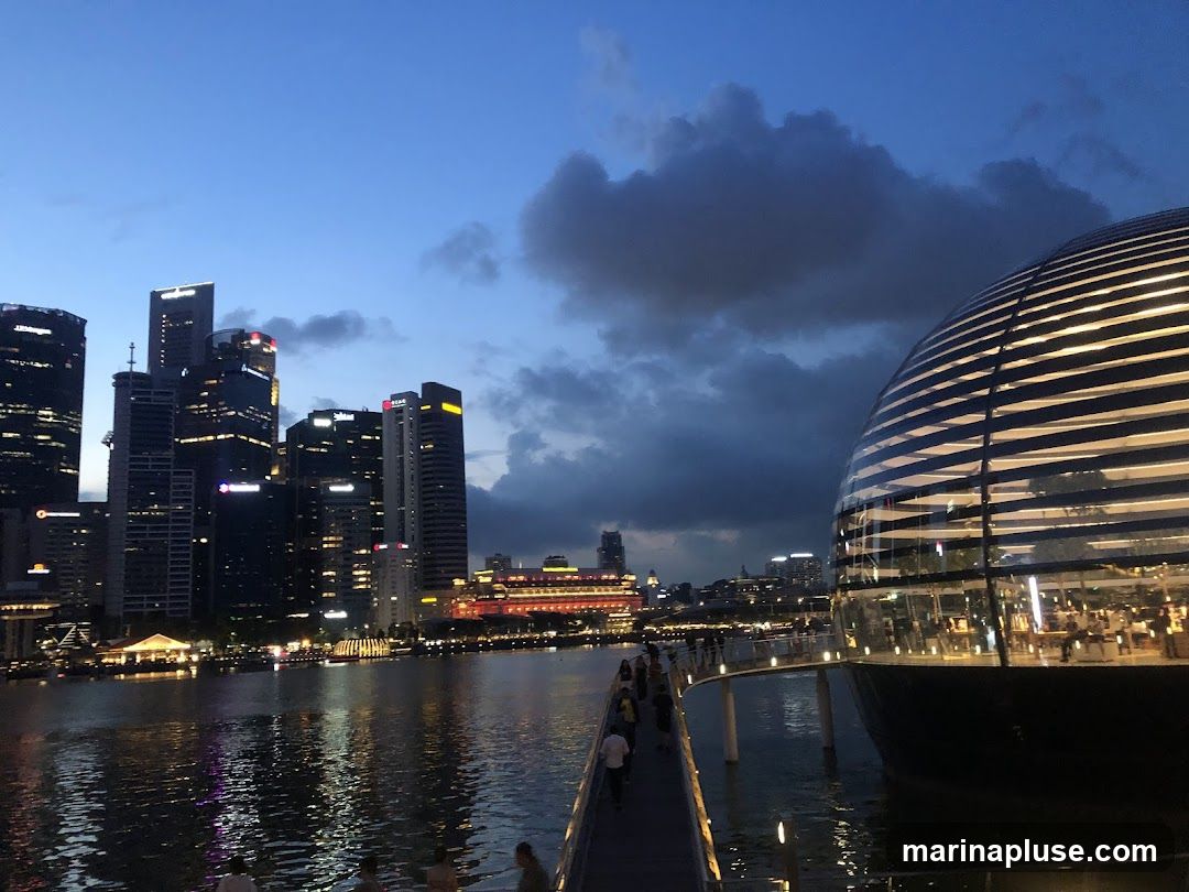 A wide shot of a modern cityscape at dusk, with a body of water in the foreground reflecting the lights of the buildings. A distinctive spherical glass building with illuminated horizontal stripes is prominent on the right. A walkway leads towards it from the left, with several people visible. In the background, a cluster of tall skyscrapers are lit up against the darkening blue sky, with some clouds present.