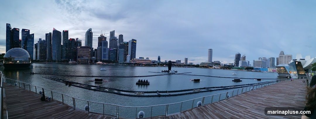 A panoramic view of the Marina Bay skyline in Singapore during dusk. The image captures the modern architecture of the city's skyscrapers, a domed building on the water, and the distinctive lotus-shaped ArtScience Museum. Floating structures and a light show setup are visible in the bay. The foreground features a wooden boardwalk with railings, and a few people are present. The sky is overcast with a soft, diffused light.