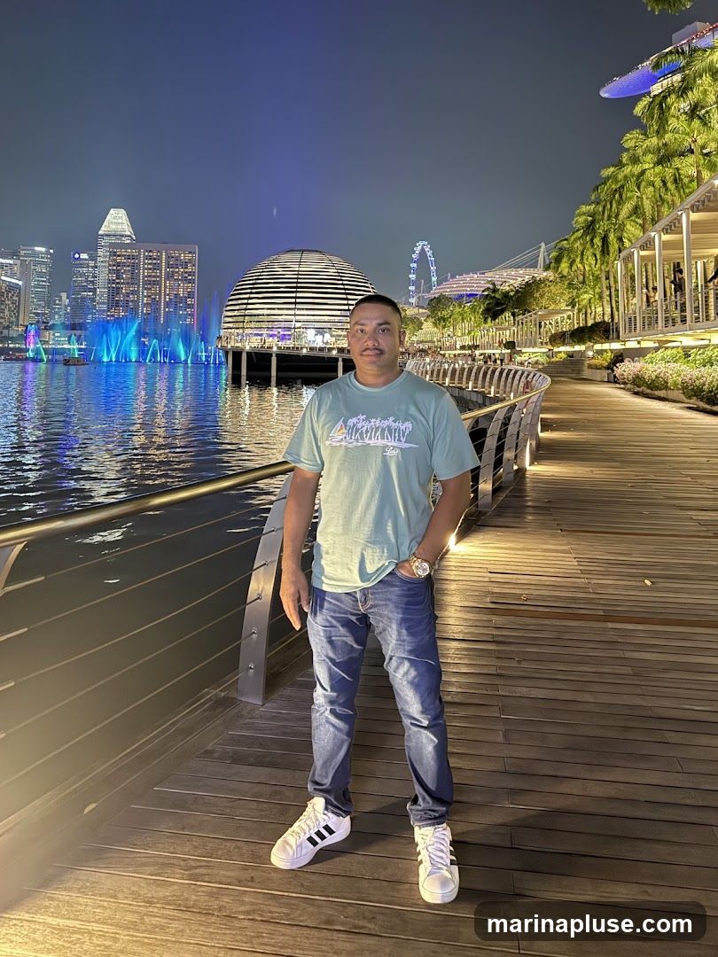 A man stands on a wooden boardwalk at night, with a city skyline and a Ferris wheel in the background. The city lights reflect on the water, and there are fountains spraying blue lights. Palm trees line the right side of the boardwalk. The man is wearing a light green t-shirt, blue jeans, and white sneakers. He has his left hand in his pocket and is looking towards the camera.
