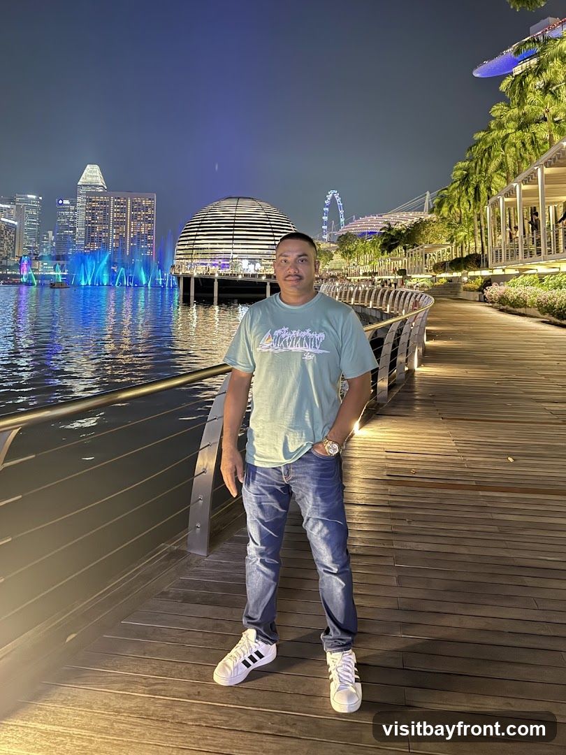 A man stands on a wooden boardwalk at night, with a city skyline and a Ferris wheel in the background. The city lights reflect on the water, and there are fountains spraying blue lights. Palm trees line the right side of the boardwalk. The man is wearing a light green t-shirt, blue jeans, and white sneakers. He has his left hand in his pocket and is looking towards the camera.