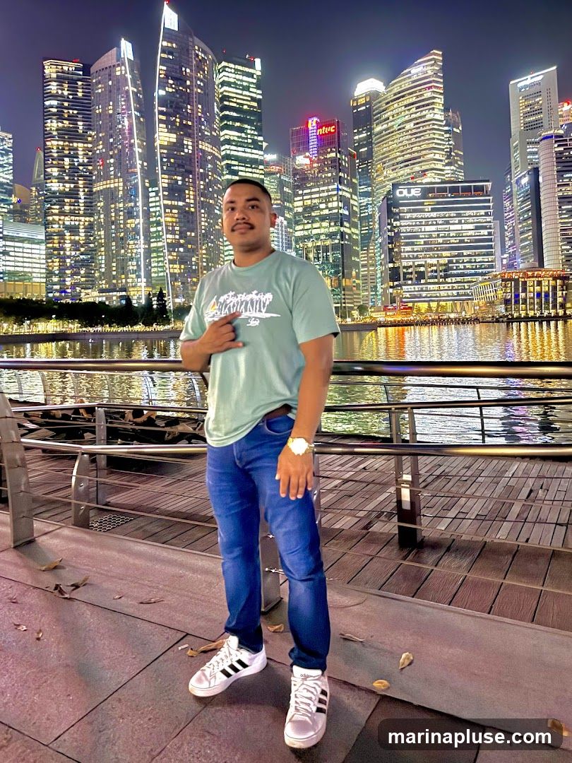 A man stands on a walkway at night, with a brightly lit city skyline and waterfront in the background. He is wearing a light green t-shirt with a graphic print, blue jeans, and white sneakers. The city buildings are illuminated with many lights, reflecting on the water.