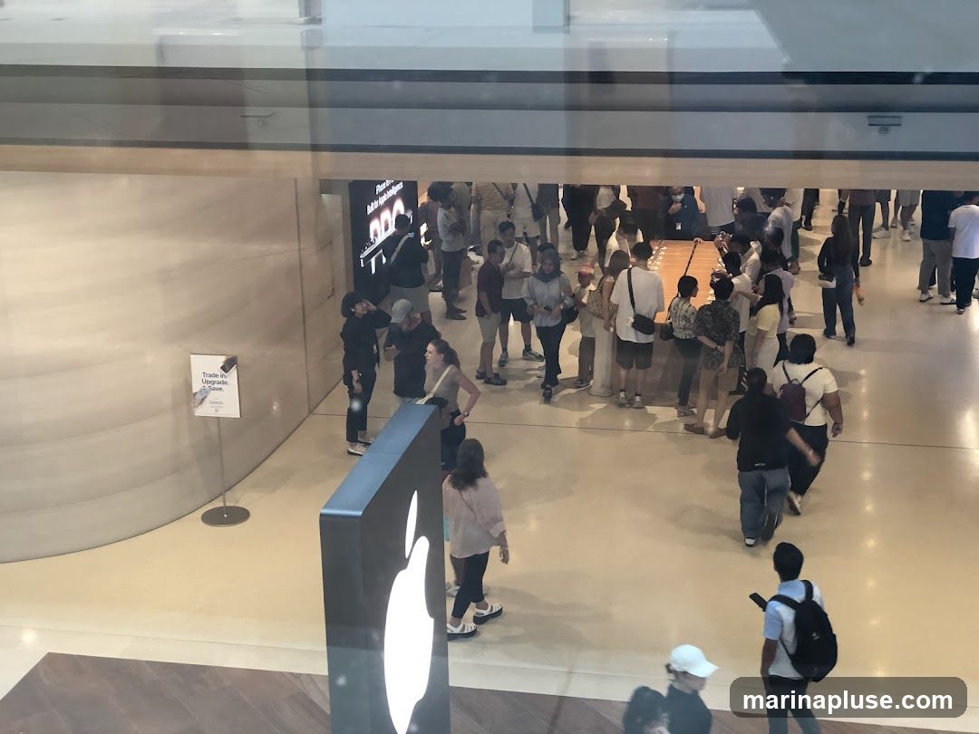 A crowd of people are gathered inside a store, with some looking at displays and others interacting with staff. There is a large sign with a logo visible in the foreground. Another sign on the wall reads "Trade in. Upgrade. Save.".