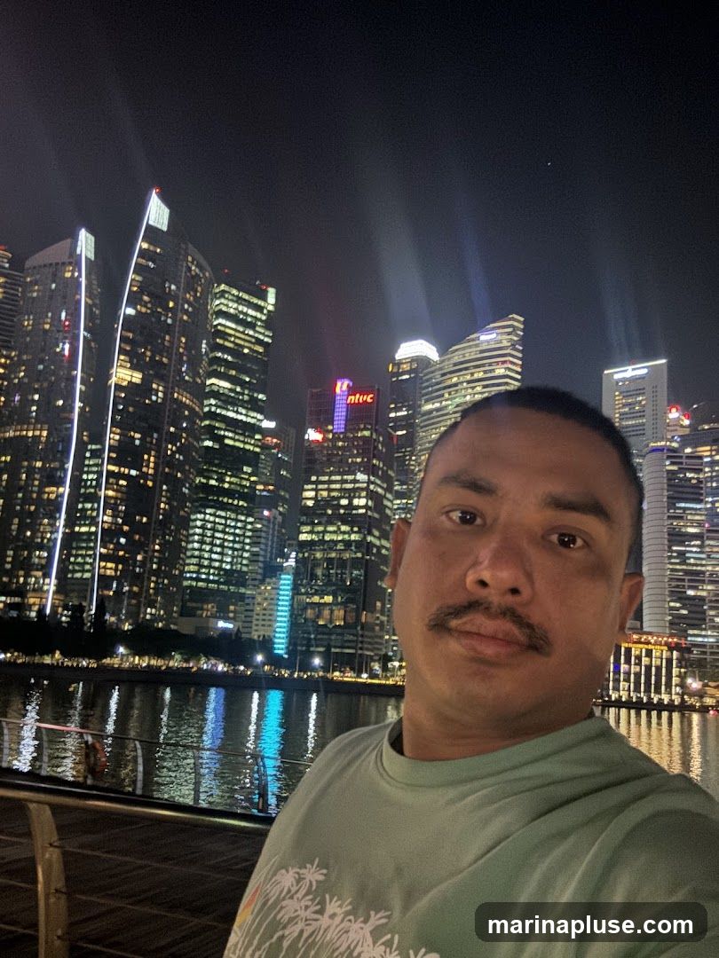 A man with a mustache is taking a selfie at night. Behind him, a city skyline is illuminated with many lights. The lights reflect on the water in front of the buildings. The man is wearing a light green t-shirt with a palm tree design.