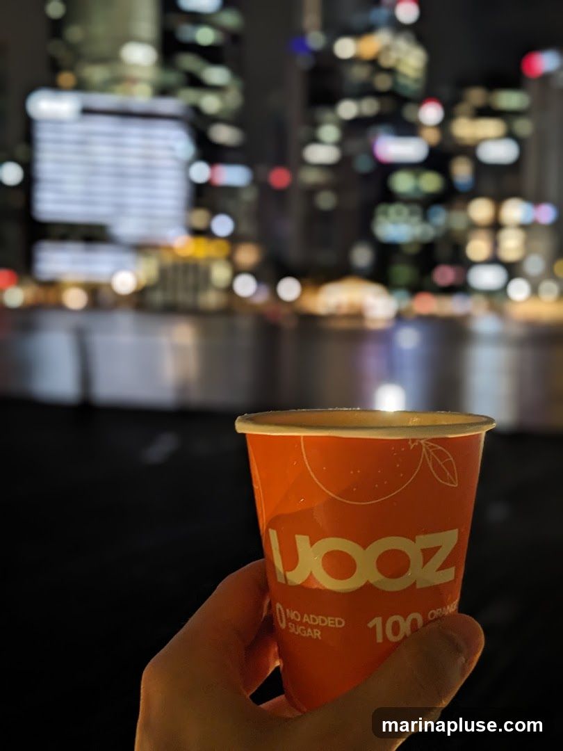 A hand holds an orange paper cup with the logo "UOOZ" and text "NO ADDED SUGAR 100 ORANGE". The background is blurred, showing city lights at night, possibly reflecting on water.