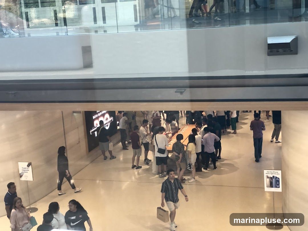 A high-angle view shows a busy store interior with many people browsing and queuing. A large screen displays text and graphics. Several display tables are visible, with customers interacting with products. Signage indicating "Trade in, Upgrade now" and "Get the iPhone that's right for you" are placed around the store.