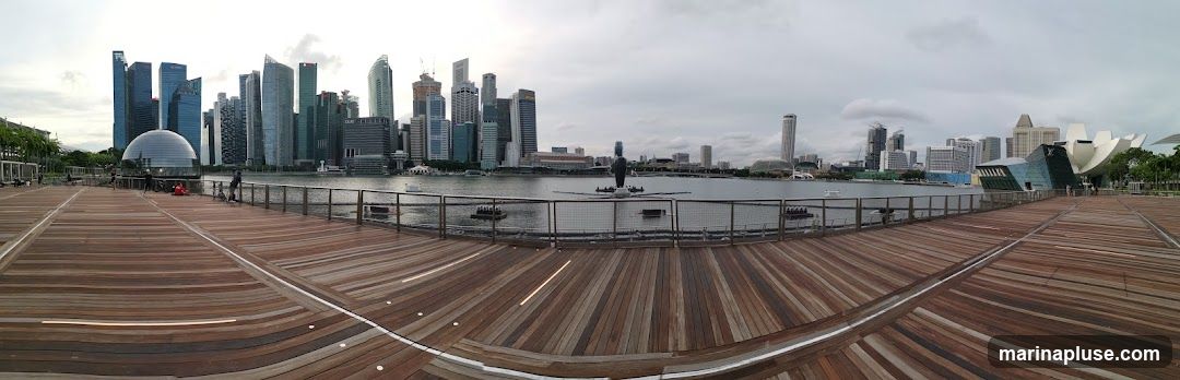 A panoramic view of the Singapore skyline, featuring the Marina Bay Sands hotel and other skyscrapers, across a body of water. The foreground is a wooden boardwalk with a curved design.