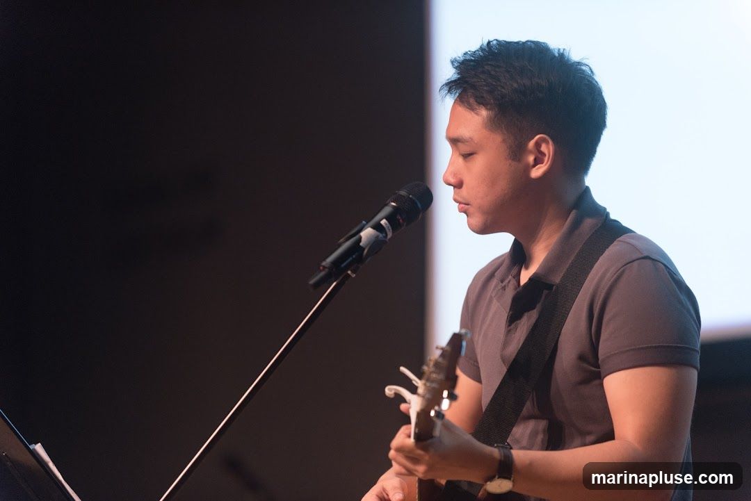 A man in a grey polo shirt plays an acoustic guitar and sings into a microphone on a stage. He is wearing a watch on his left wrist. The background is dark with a bright screen or light source on the right.