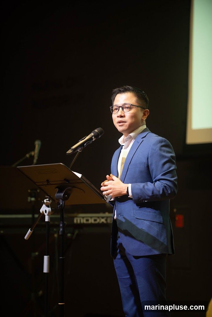 A man in a blue suit and glasses stands at a podium, speaking into a microphone. He has his hands clasped in front of him. Behind him, a keyboard with the word "MOOG" visible is dimly lit. The background is dark.