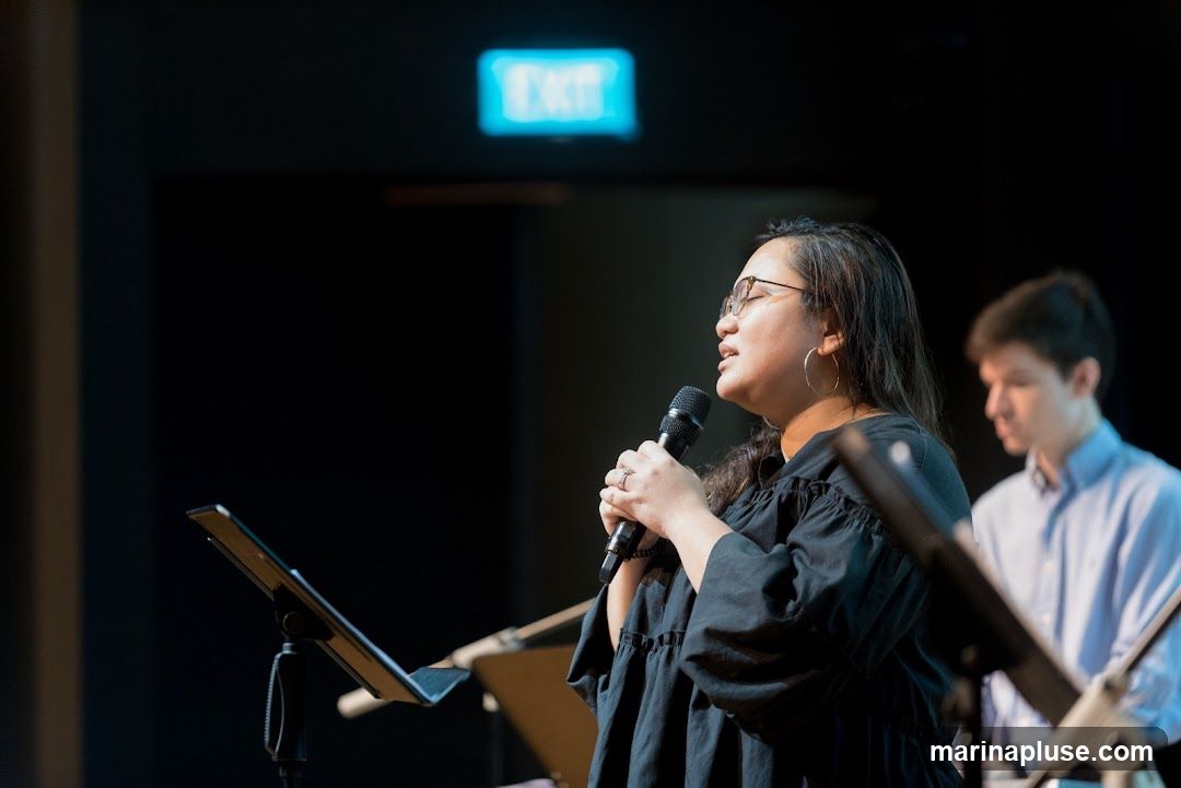 A woman wearing glasses and a black dress sings into a microphone on a stage. She is wearing hoop earrings and a ring on her left hand. In the background, a young man in a light blue collared shirt is out of focus. An illuminated blue "EXIT" sign is visible above them. There are music stands in the foreground.