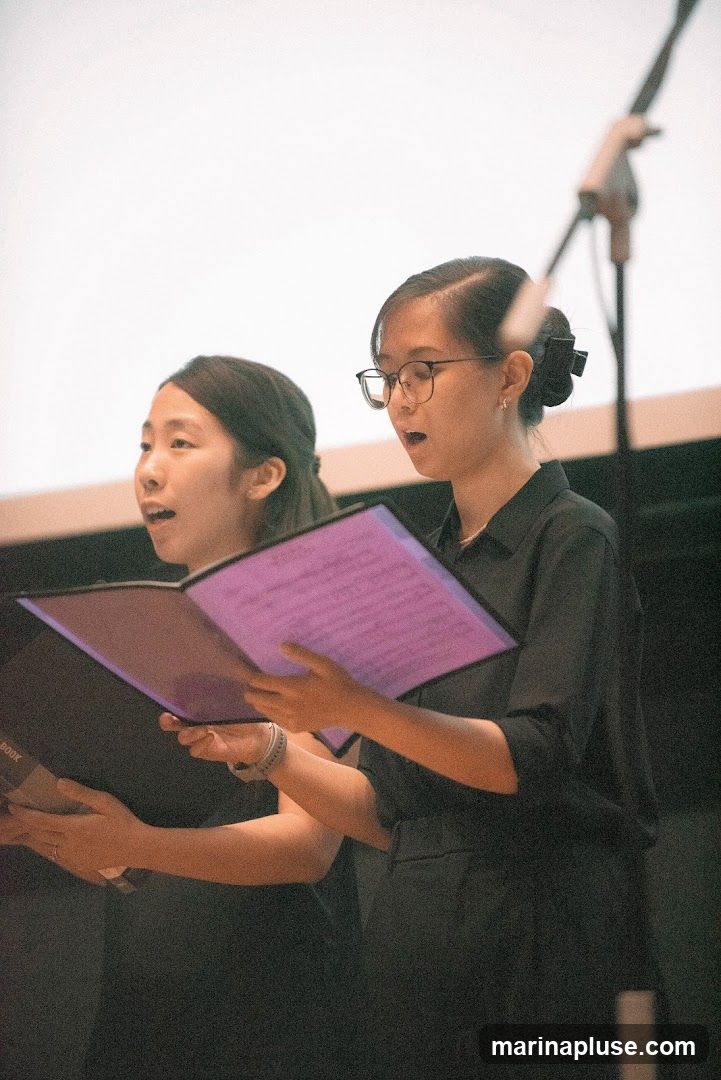 Two young women are shown from the chest up, singing from open sheet music. The woman on the right wears glasses and has her hair pulled back in a bun. The woman on the left has her hair down. Both are wearing dark clothing. A microphone stand is visible in the background.
