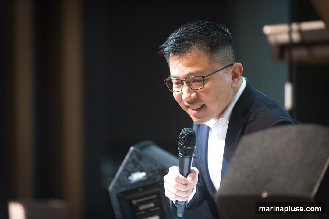 A man wearing glasses and a dark suit over a white shirt is speaking into a microphone. He is holding the microphone in his right hand and appears to be on a stage or in a presentation setting. There is a speaker to his left and a blurred background with vertical lines.