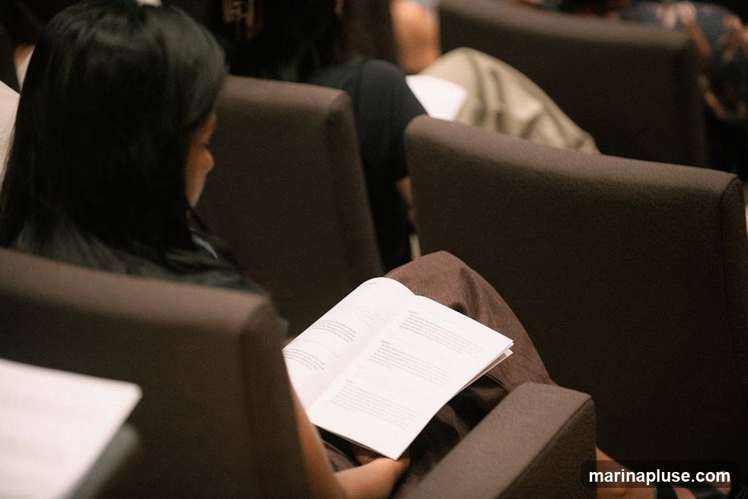 A woman with long dark hair is reading a book in a lecture hall or auditorium. She is seated in a dark brown chair, wearing brown pants. Other audience members are visible in the background, also seated in chairs.