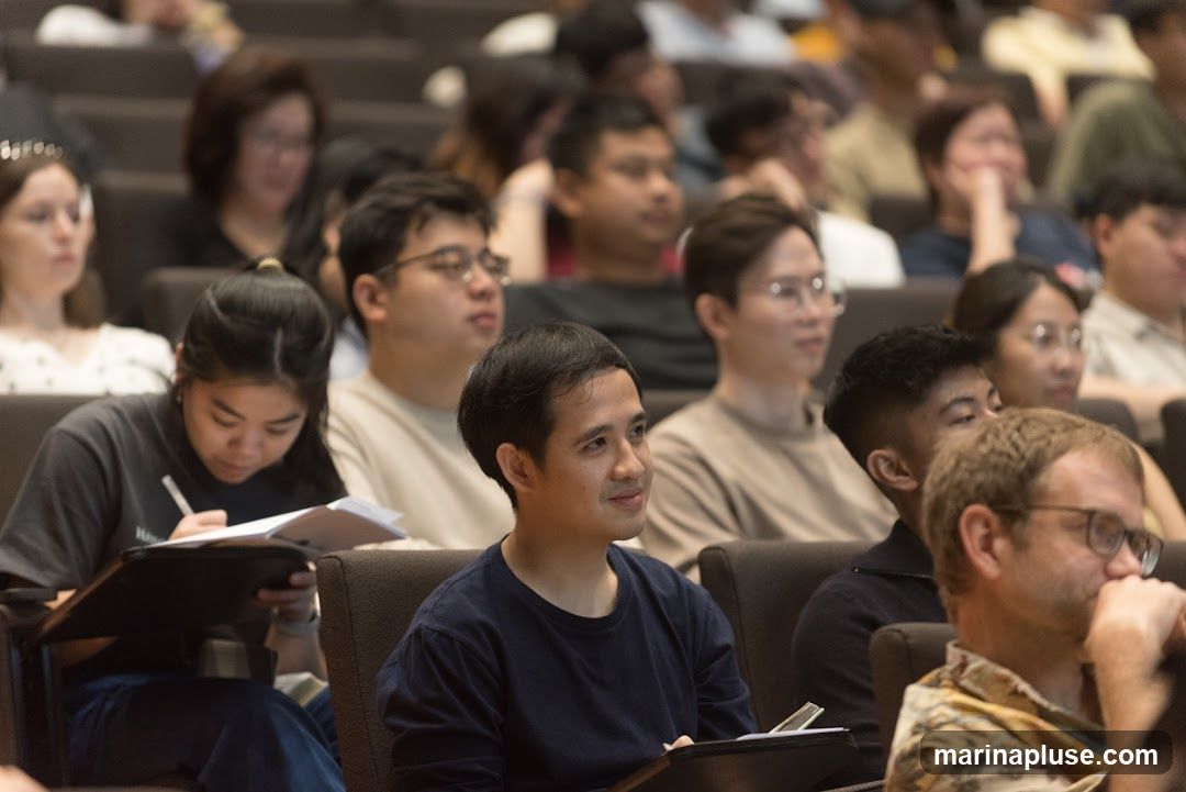 A diverse group of people are seated in an auditorium, facing forward. Some are taking notes, while others are looking ahead. The lighting suggests an indoor event, possibly a lecture, presentation, or conference.