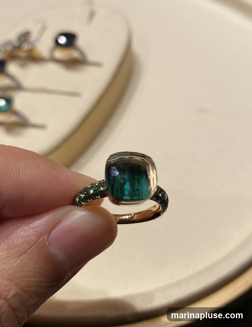 A hand holds a ring with a large, square-cut, multi-colored gemstone. The band of the ring is adorned with small green stones. In the blurred background, several other rings are displayed on a beige stand.