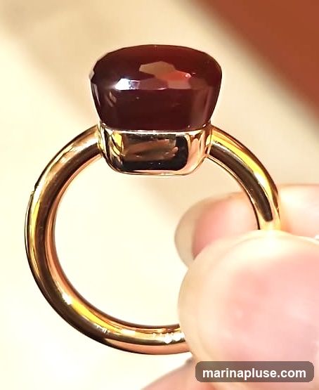 A close-up shot of a ring with a dark red, faceted gemstone. The band of the ring appears to be gold. A person's fingers are holding the ring.