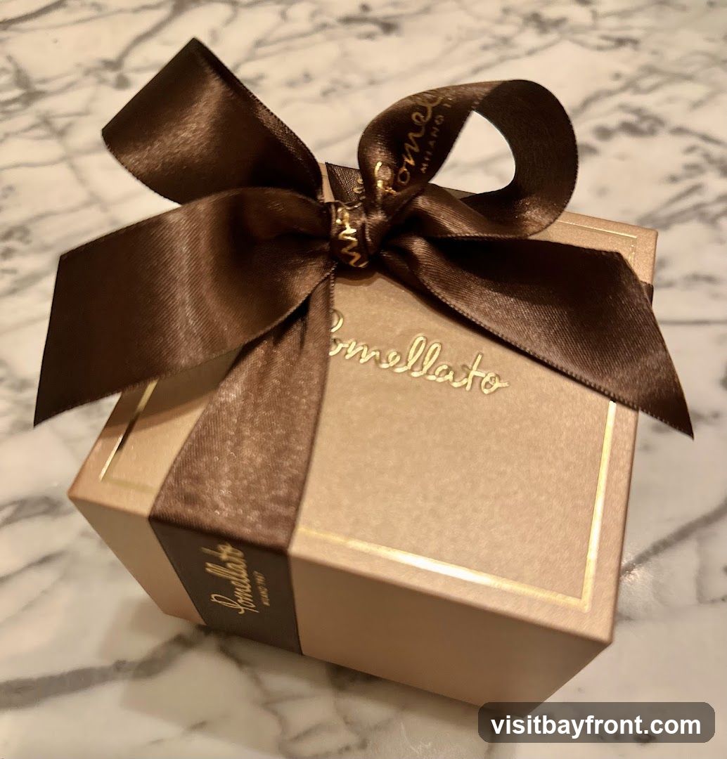 A close-up shot of a light brown Pomellato gift box tied with a dark brown satin ribbon. The Pomellato logo is embossed in gold on the front of the box and on a band around the side. The ribbon is tied in a bow on top of the box.