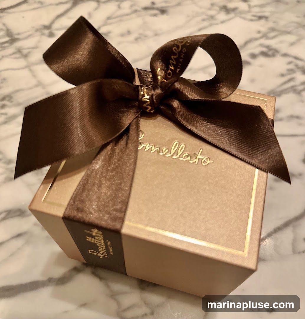 A close-up shot of a light brown Pomellato gift box tied with a dark brown satin ribbon. The Pomellato logo is embossed in gold on the front of the box and on a band around the side. The ribbon is tied in a bow on top of the box.