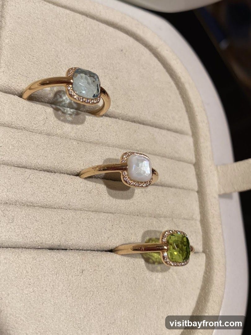 Three gold rings are displayed on a beige velvet ring holder. The top ring features a light blue gemstone surrounded by small diamonds. The middle ring has a pearlescent white gemstone, also encircled by small diamonds. The bottom ring showcases a vibrant green gemstone, similarly accented with diamonds.