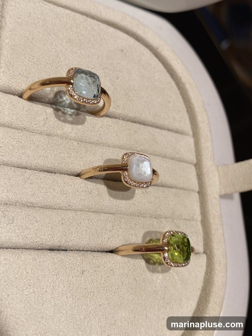 Three gold rings are displayed on a beige velvet ring holder. The top ring features a light blue gemstone surrounded by small diamonds. The middle ring has a pearlescent white gemstone, also encircled by small diamonds. The bottom ring showcases a vibrant green gemstone, similarly accented with diamonds.