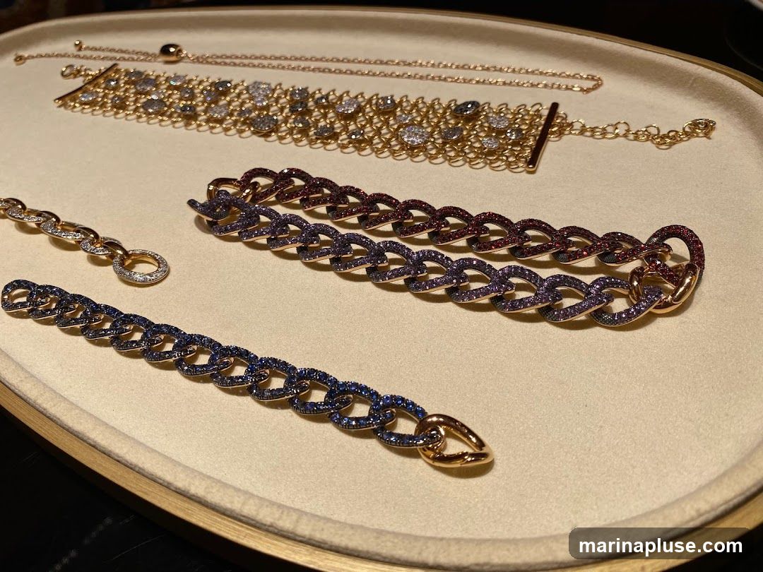 A close-up shot displays four bracelets and a delicate necklace arranged on a beige tray. The top bracelet is a wide, gold-toned band adorned with numerous clear and grey gemstones. Below it, two curb-link bracelets are featured: one with red and purple gemstones, and another with blue gemstones. To the left, a smaller rose gold bracelet with diamond accents is visible. A thin rose gold necklace with an adjustable clasp lies across the top of the tray.