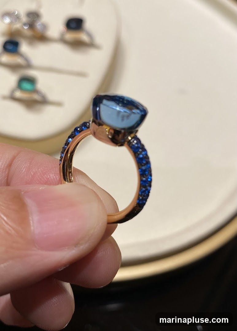 A hand holds a rose gold ring featuring a large, faceted blue gemstone. The band of the ring is encrusted with small blue stones. In the blurred background, several other rings are displayed on a white surface.