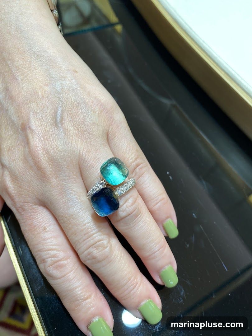 A hand wearing a ring with two large, square-cut gemstones, one blue and one teal, set in a band encrusted with small diamonds. The hand has light green nail polish. The background is dark and reflective, suggesting a display case or polished surface.