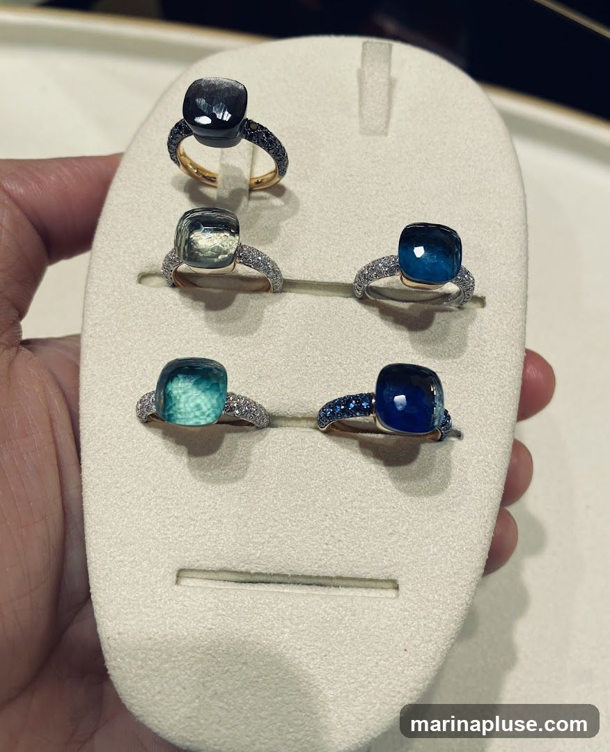 A hand holds a display stand showcasing five rings, each featuring a large, cushion-cut gemstone. The gemstones vary in color, including dark gray, light green, teal, and two shades of blue. The rings are set in gold and silver bands, some adorned with smaller diamonds or blue stones. The display stand is a neutral beige color, designed to highlight the jewelry.