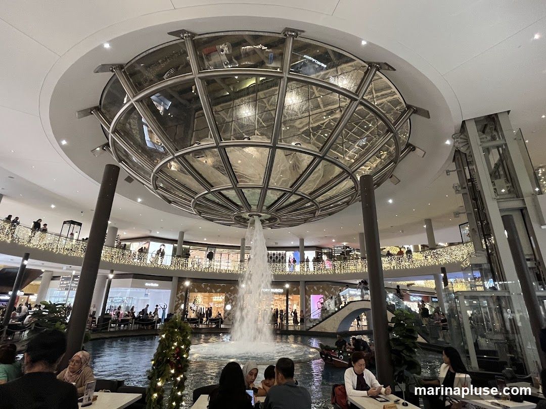 An indoor shopping mall features a large, multi-level water fountain at its center, surrounded by a canal. People are seated at tables in a dining area overlooking the water. Shops and escalators are visible on the upper levels, adorned with festive lights.