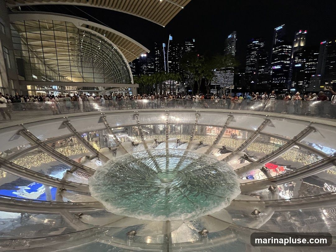 A large, circular fountain with swirling water is in the foreground, illuminated by lights. Behind the fountain, a modern building with a glass facade is visible, with a crowd of people gathered in front of it. In the background, a city skyline with illuminated skyscrapers stands against the night sky.