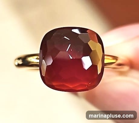 A close-up shot of a ring featuring a large, cushion-cut red gemstone. The gemstone is faceted, creating many reflective surfaces that catch the light. The band of the ring appears to be gold. The background is softly blurred, with a warm, neutral tone.
