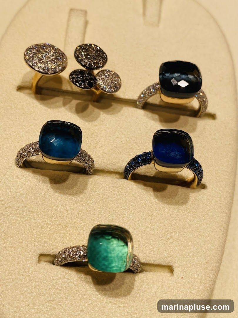 A display of several rings featuring large, cushion-cut gemstones in shades of deep blue, teal, and green, set in gold bands encrusted with small diamonds. Two rings in the background have circular, pave-set diamond and black stone tops. The rings are presented on a beige, textured display stand.