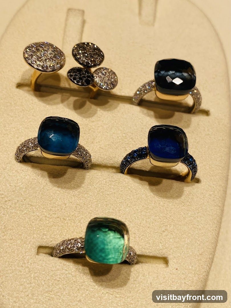 A display of several rings featuring large, cushion-cut gemstones in shades of deep blue, teal, and green, set in gold bands encrusted with small diamonds. Two rings in the background have circular, pave-set diamond and black stone tops. The rings are presented on a beige, textured display stand.